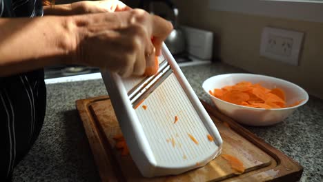 Woman-Hands-Shredding-Carrot-On-A-Manual-Grater-On-Top-Of-A-Chopping-Board-In-The-Kitchen---closeup