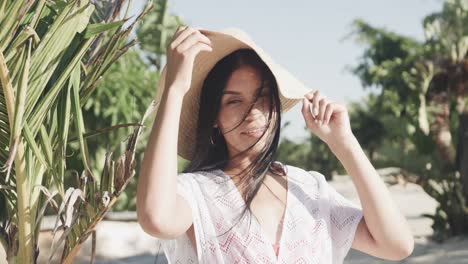 portrait of happy hispanic woman in sunhat and sundress smiling on sunny beach, slow motion