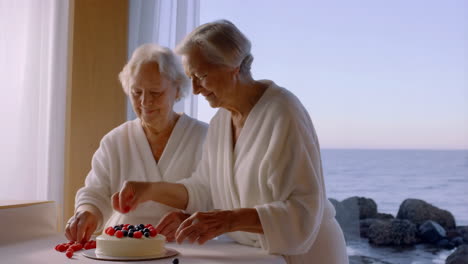 two senior women in bathrobes decorating a cake with berries by a window overlooking the ocean