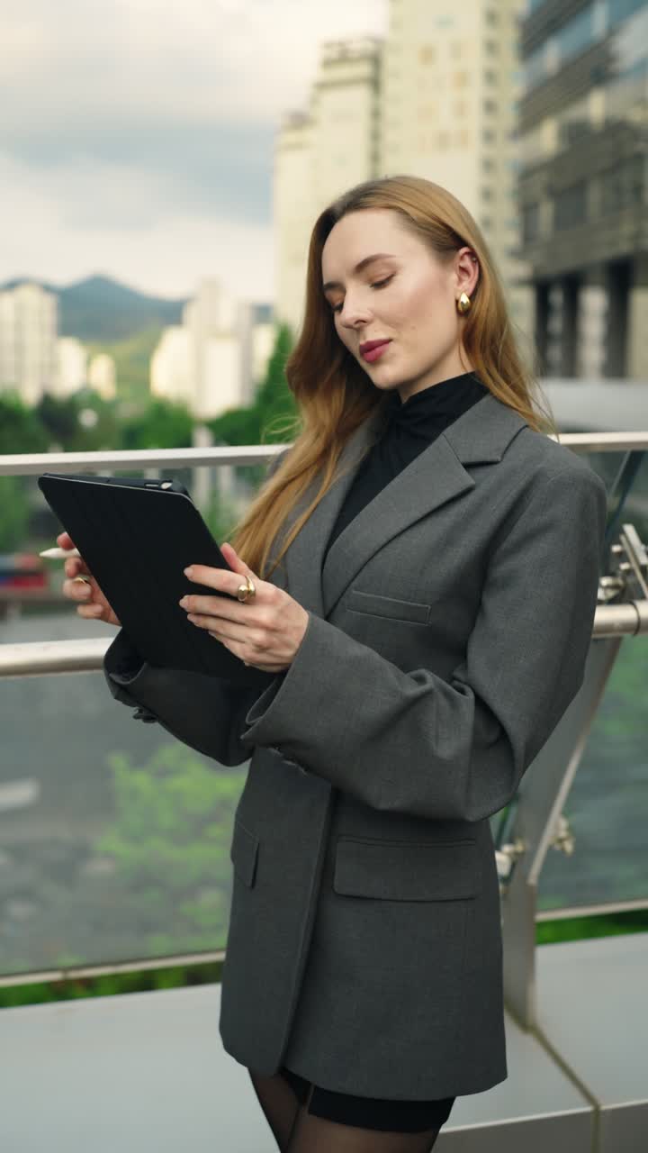 Caucasian businesswoman in a gray suit stands outdoors by office buildings, thinking with a digital tablet and pen, then writing notes in a vibrant city setting