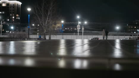 nighttime view of two people walking near an illuminated urban area, with streetlights and passing cars in the background