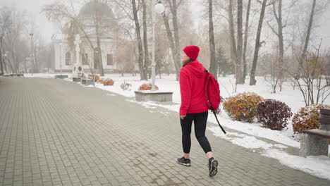 woman with bag over shoulder walking along interlocked pathway surrounded by trees, benches, light poles, and snow-covered ground in foggy atmosphere with distant building visible