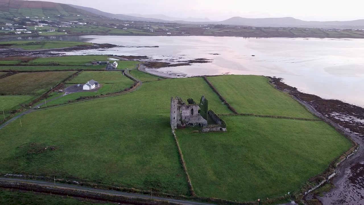 Drone establishing shot capturing the historic ruins of Ballycarbery Castle situated in vibrant green pastures alongside a calm coastal estuary in rural Kerry, Ireland
