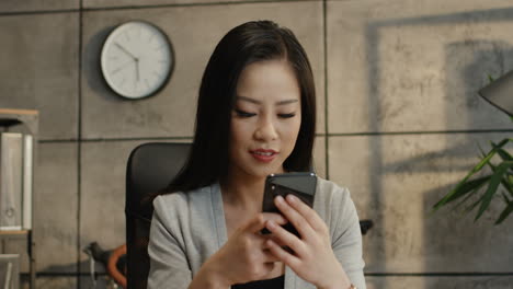 Close-Up-Of-The-Young-Pretty-Smiled-Businesswoman-Reading-Something-On-The-Smartphone-Screen-And-Texting-On-It-In-The-Office-And-Then-Looking-At-The-Side