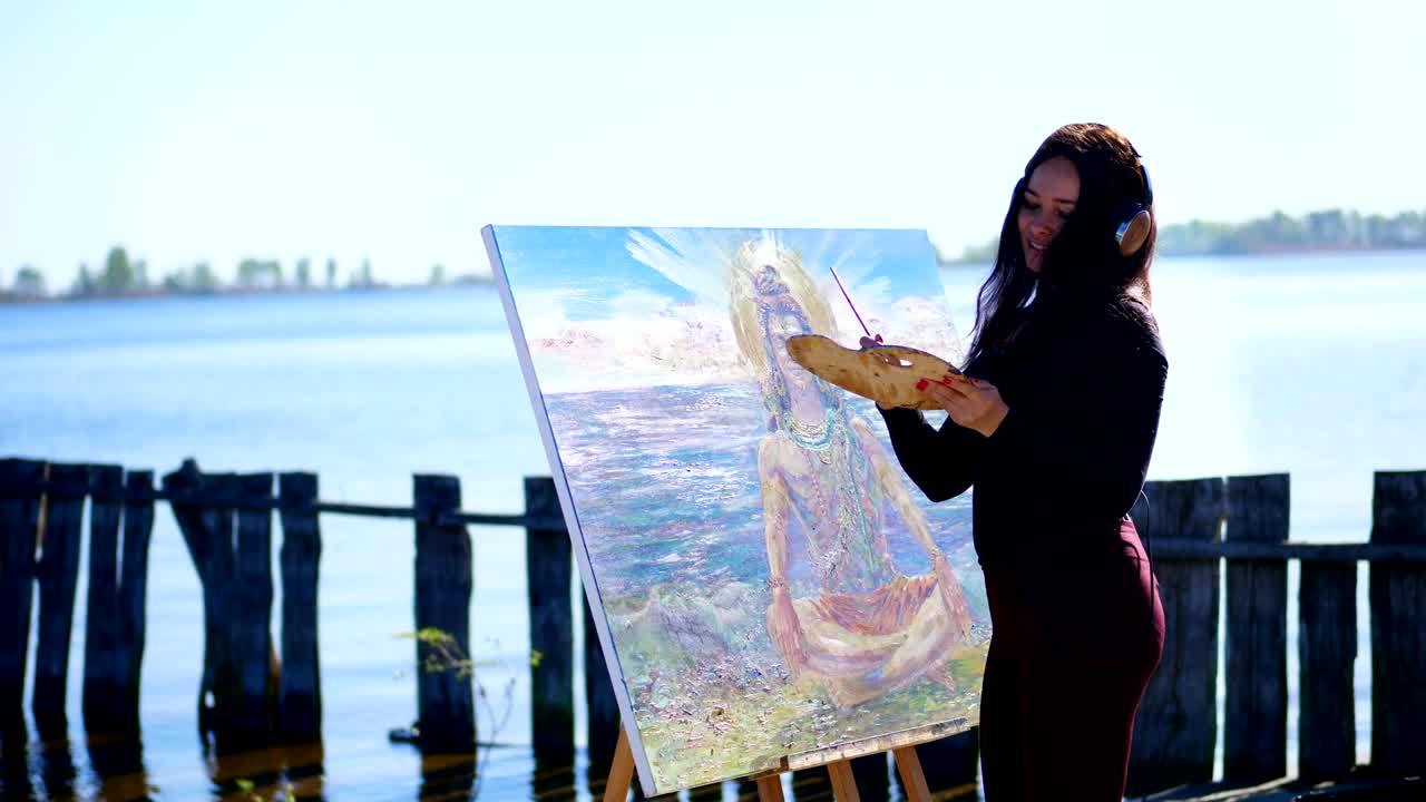 여름, 강 근처, 해변. 해가 뜨면, 헤드폰을 입은 아름다운 여성 예술가는 을 들고, 손에 팔레트를 들고, 캔버스 신 시바에 그림을 그리고, 음악을 듣고, 휴식을 취하고, 춤을 춘다.