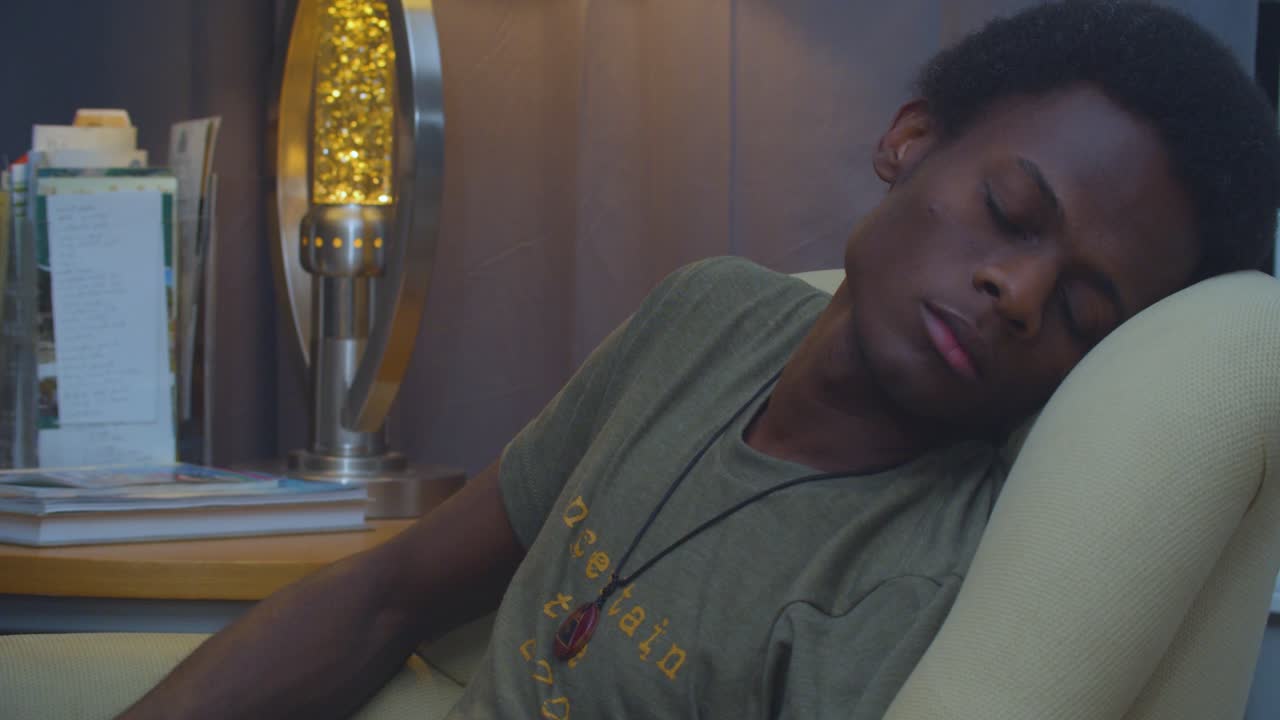 A slow motion and cinematic eye level static shot of a tired, yet handsome skinny young black-African American man napping in the living room with a lamp in the background