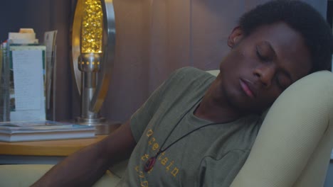A-slow-motion-and-cinematic-eye-level-static-shot-of-a-tired,-yet-handsome-skinny-young-black-African-American-man-napping-in-the-living-room-with-a-lamp-in-the-background