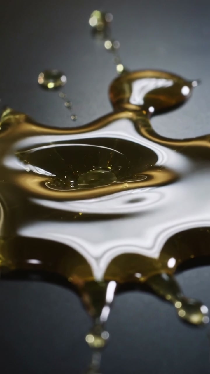 Vertical video: Falling droplet striking amber viscous puddle on dark tabletop, forming bubbles