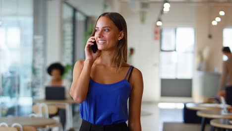 Young-Smiling-Businesswoman-Standing-In-Busy-Modern-Open-Plan-Office-Talking-On-Mobile-Phone