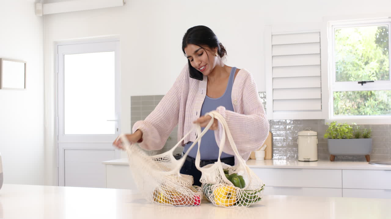 Woman in kitchen talking on phone while unpacking groceries from reusable bags