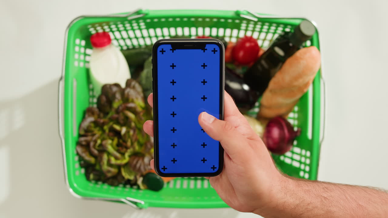 Person holding a smartphone with blue screen, over a grocery basket filled with food