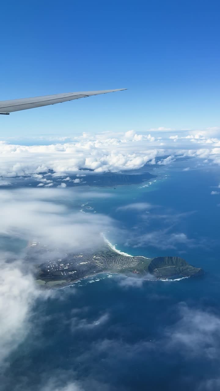 Pemandangan dari pesawat yang menunjukkan sayap, awan, lautan, dan Pulau Oahu di bawah.