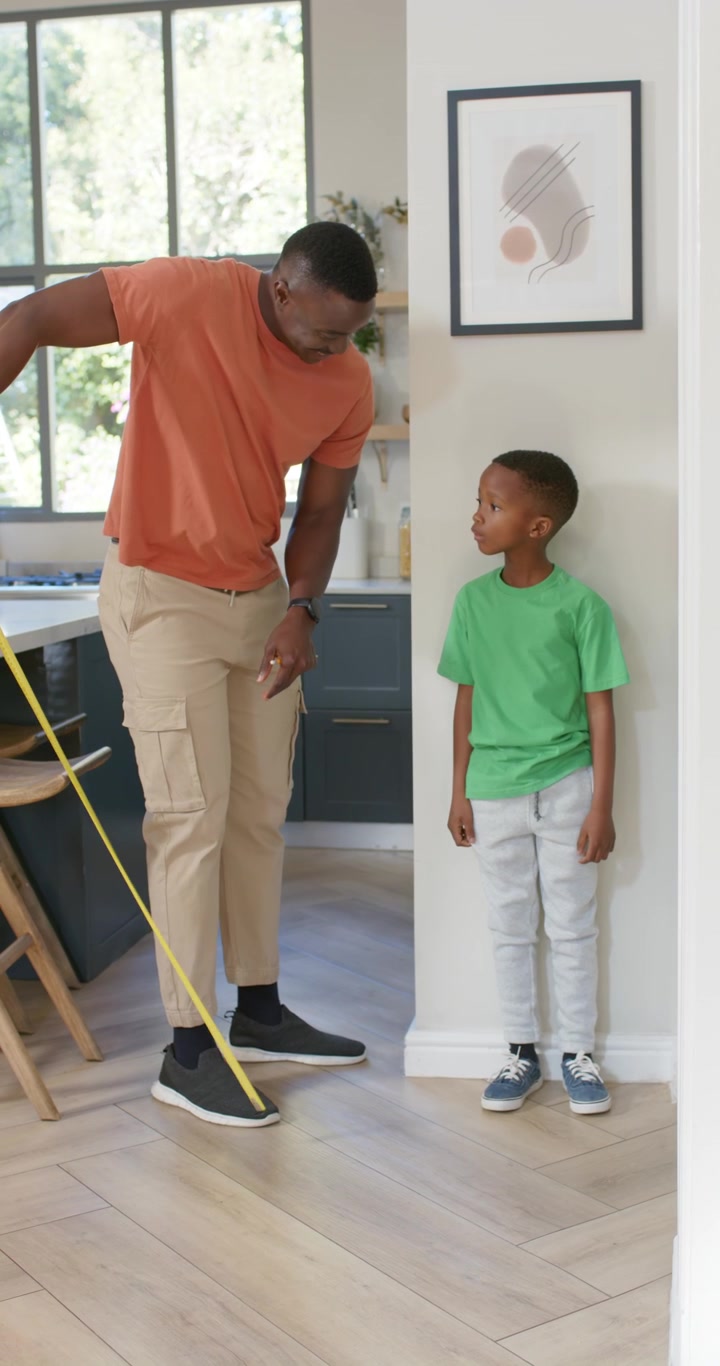 Vertical video: African American dad and son in hall measuring height, pulling yellow tape to floor