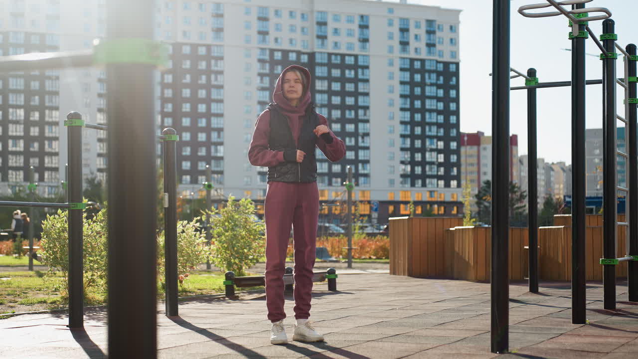 Jogger wearing hoodie and vest stretches one arm across chest while balancing in outdoor urban park with playground bars and pedestrians walking under autumn sunlight for workout warm up routine