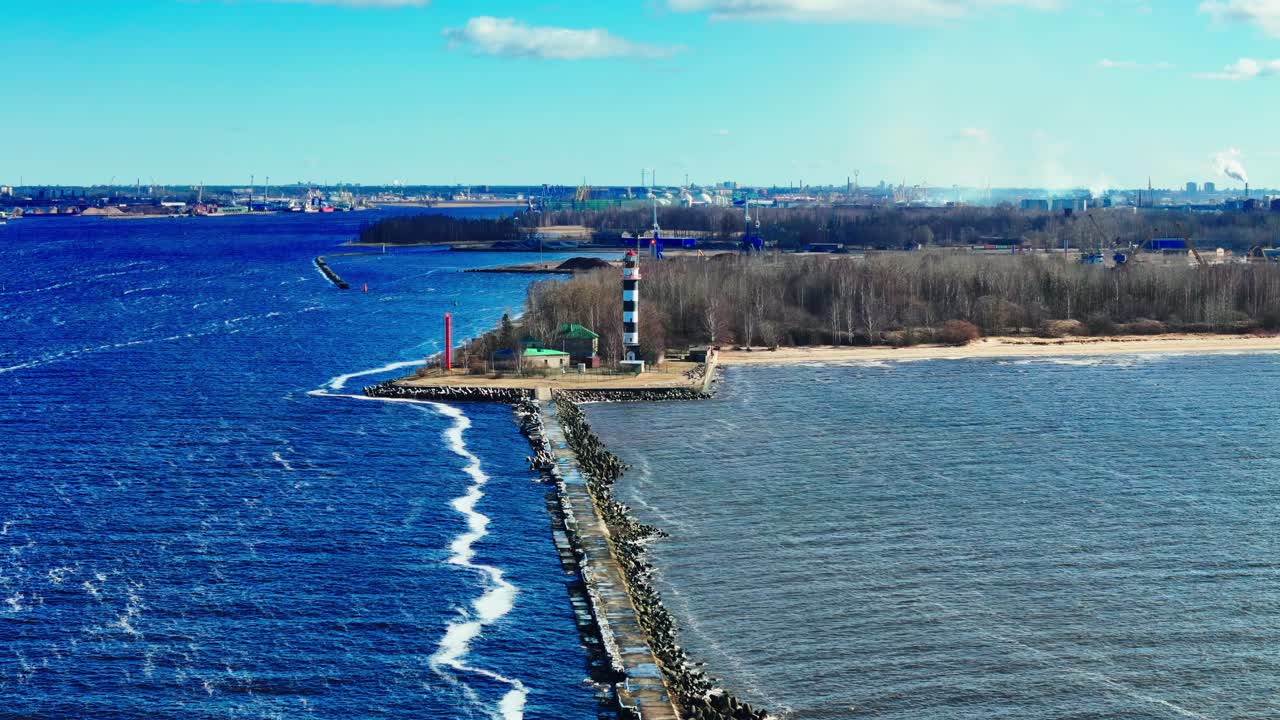 A tall striped lighthouse anchors an extended rocky breakwater, surrounded by bright blue waves and a quiet bare shoreline under open skies.