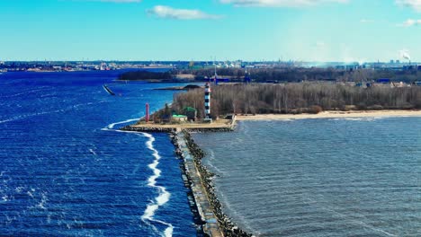 a tall striped lighthouse anchors an extended rocky breakwater, surrounded by bright blue waves and a quiet bare shoreline under open skies.