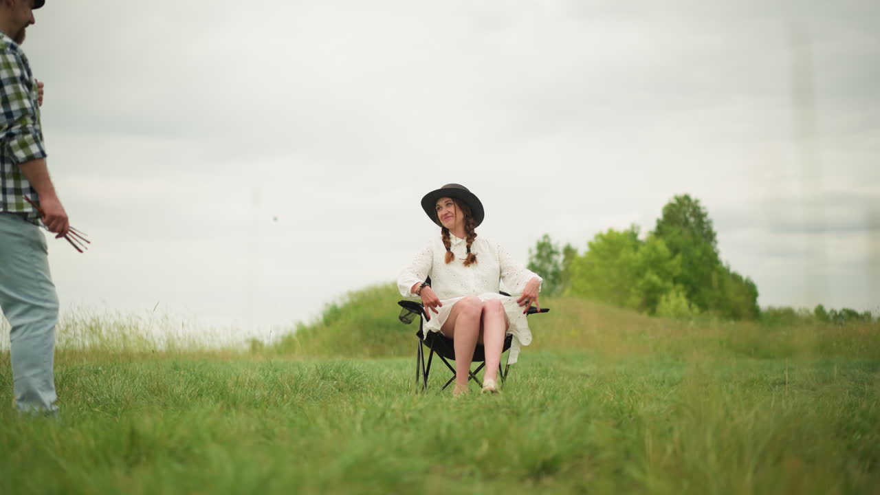 색 드레스와 검은색 모자를 입은 아름다운 여성이 잔디에 있는 의자에 앉아 자신감 있게 포즈를 취하고 있다. 색 셔츠를 입은 남자가 을 들고 그녀를 면밀히 지켜보고 있다.