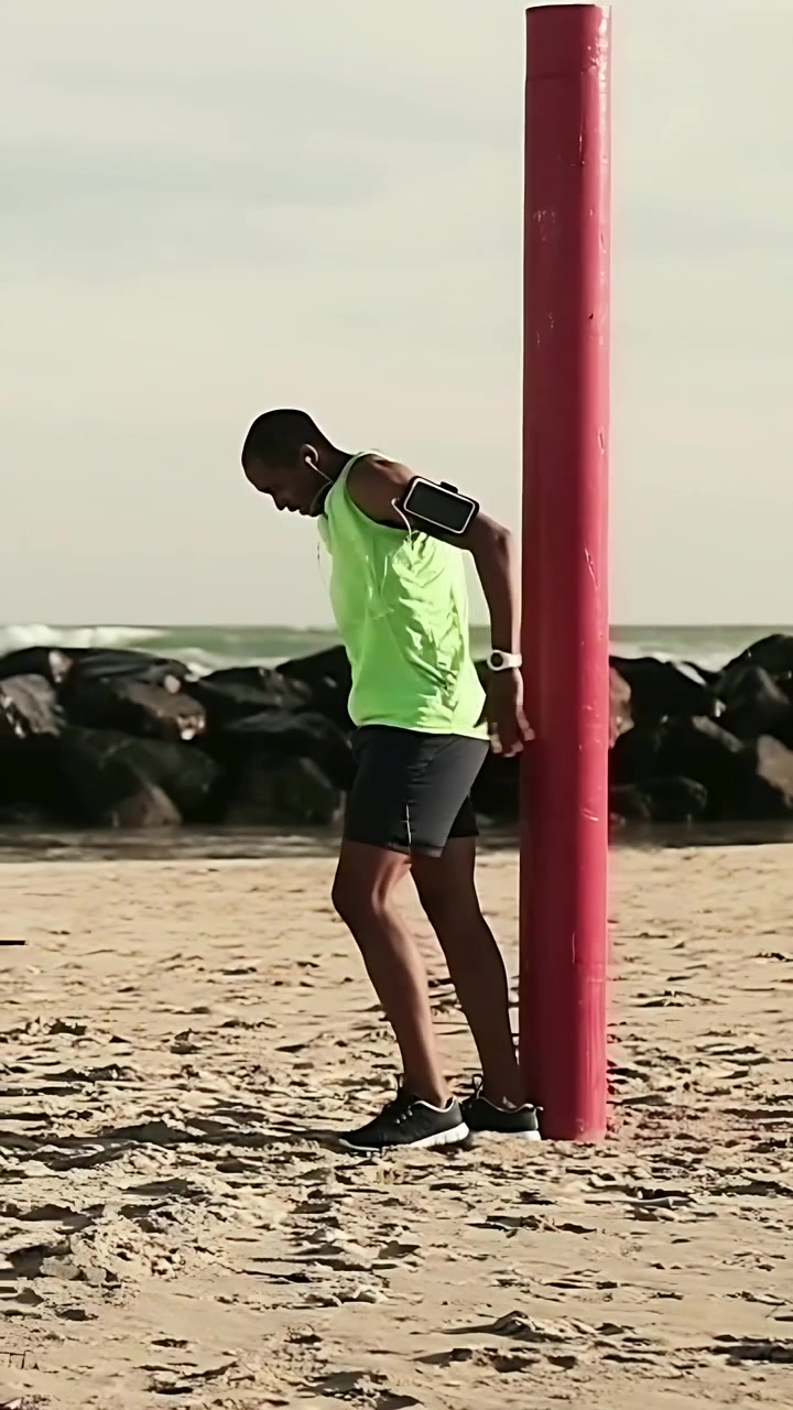 Vertikal video: Skyver fra rød stolpe, mann spurter over strand i neonfarget topp, armbånd for trening