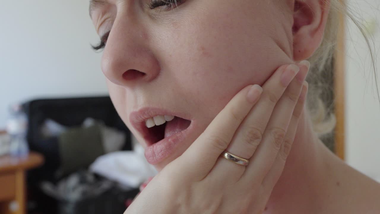 Close-up of a woman holding her cheek and reacting to tooth pain. The facial expression and hand gesture indicate discomfort, possibly from dental or gum issues
