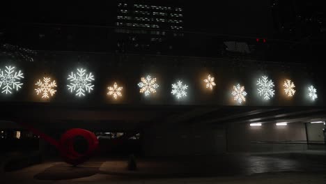 Una-Colección-De-Brillantes-Adornos-Navideños-Con-Copos-De-Nieve-Cubre-El-Costado-De-Un-Puente-En-Una-Ciudad-Con-Poca-Luz-Por-La-Noche-Con-Diferentes-Tamaños-Y-Colores