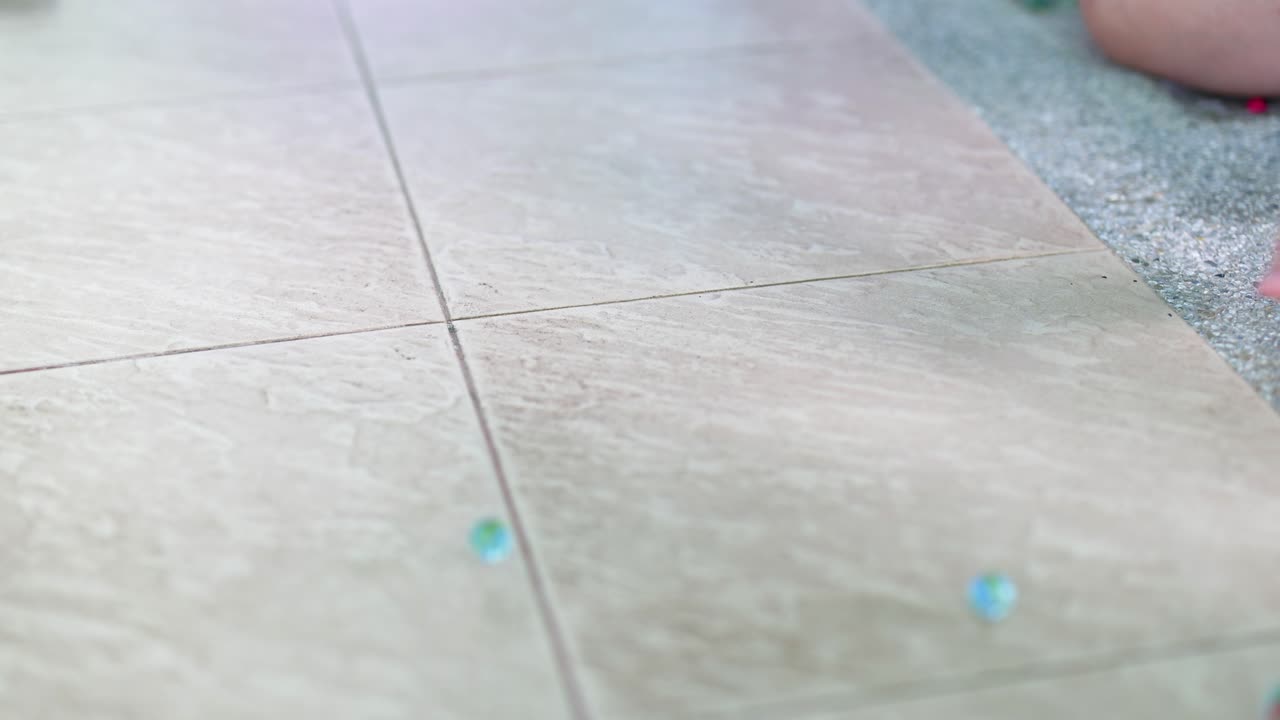 A hand releases marbles onto a tiled floor, capturing their movement and scattering. Soft lighting creates a calm atmosphere