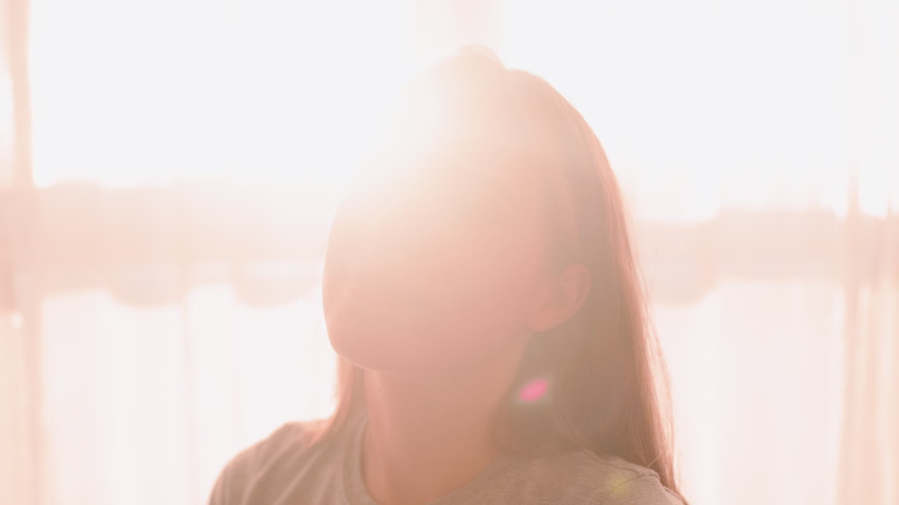 Close up of girl posing with elegance in soft morning sunlight, gentle smile on face, warm natural glow highlighting features as light flare surrounds her in serene indoor atmosphere