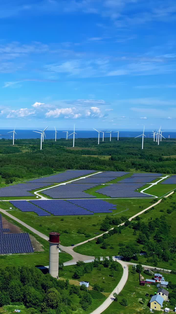 Stunning aerial shot of the Tārgale Solar Park and wind farm, showing photovoltaic panels and turbines producing sustainable energy with the Baltic Sea and a landmark water tower in view.
