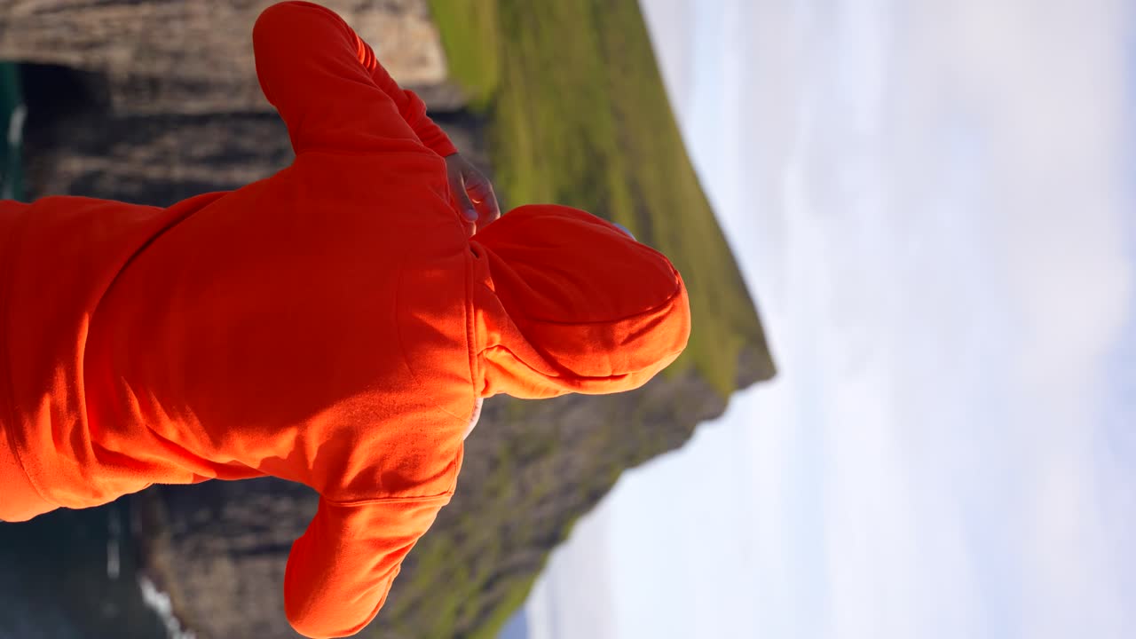 Young man in cap facing green cliff puts on hood of orange sweatshirt