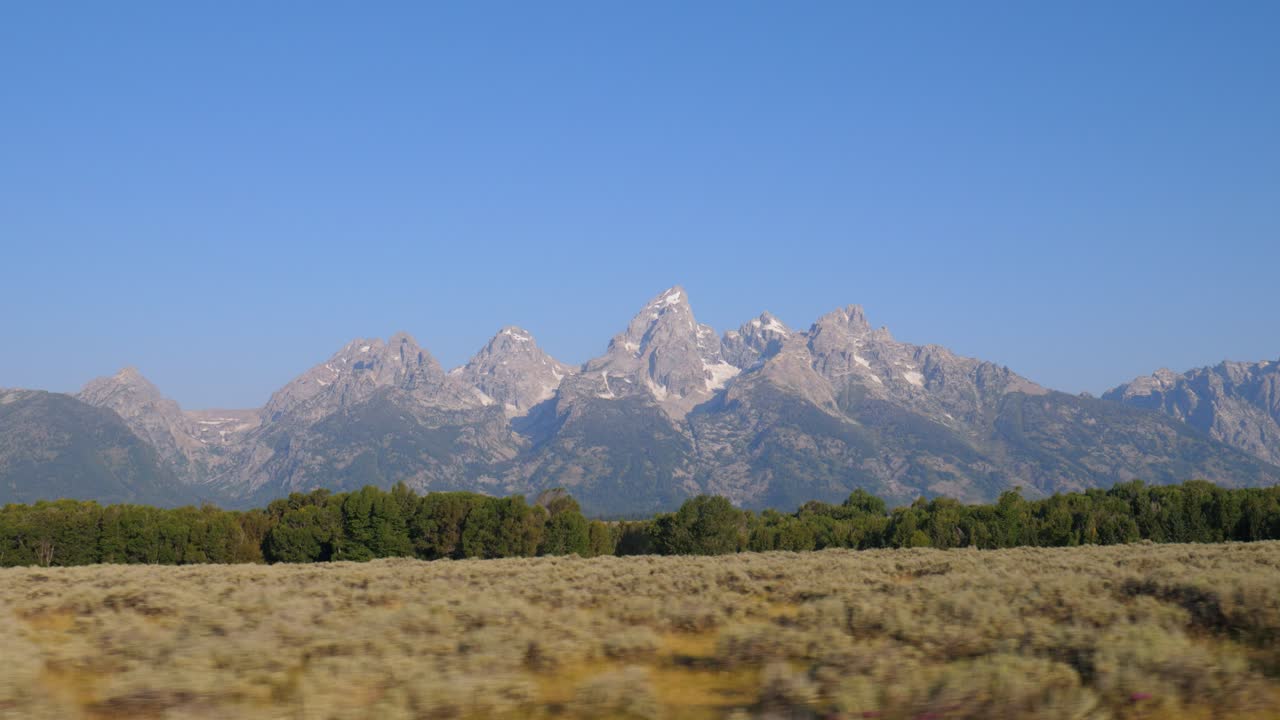 Majestic peaks of the Teton Range rise above forested slopes and golden grasslands in Grand Teton National Park. A crisp, clear view of Wyoming’s rugged alpine beauty under blue skies