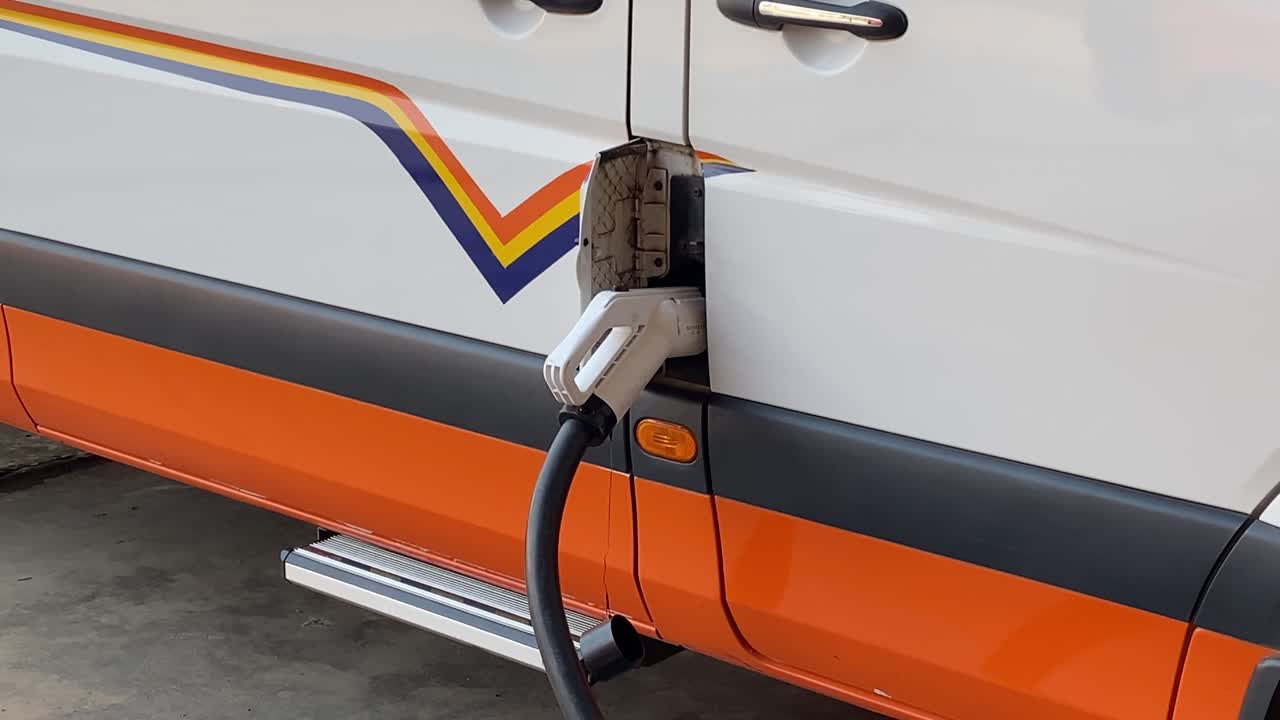 Furgone elettrico in carica presso una stazione, primo piano sul cavo collegato e sul pannello laterale.