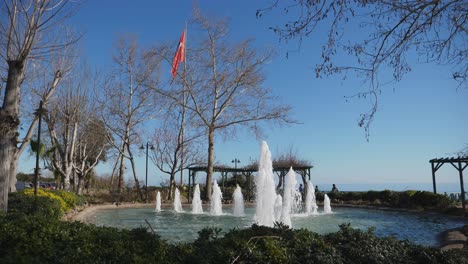 Establecimiento-De-Tiro,-Parque-De-Fuentes-En-árboles-De-Antalya-Y-Bandera-De-Pavo,-Gente-Caminando-En-El-Fondo