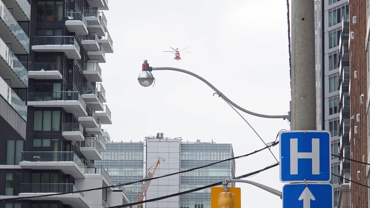 Snow falls gently on quiet downtown street with hospital sign above and lamp post along buildings framing helicopter in sky