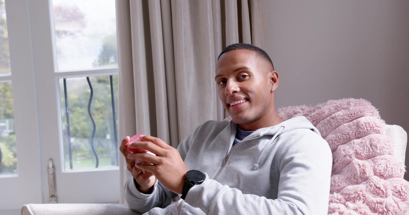 Adult African American man holding and examining pink orb, rotating in chair by window smiling