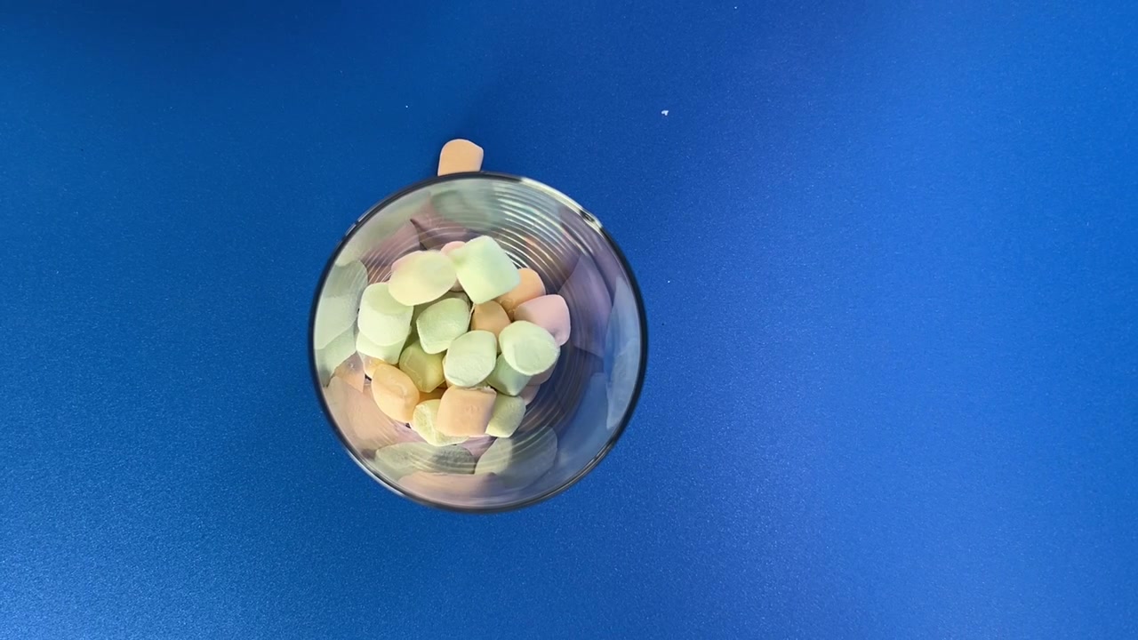 Pezzi colorati di marshmallow che cadono in una ciotola di vetro su sfondo blu, con più pezzi che cadono nell'aria durante un'azione alimentare giocosa e un movimento dinamico