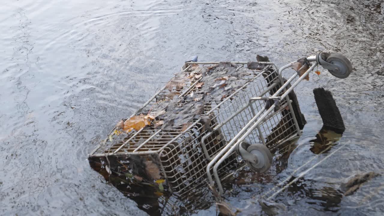 Old rusty shopping trolley laying in a lake with leaves during the day with rain, wide shot