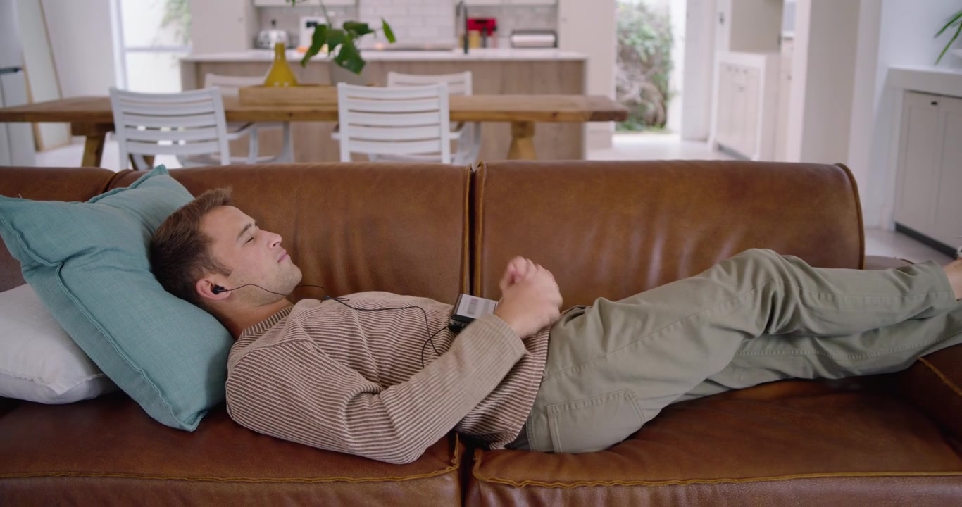 Man pressing player, starting music with wired earbuds, reclining on couch and relaxing