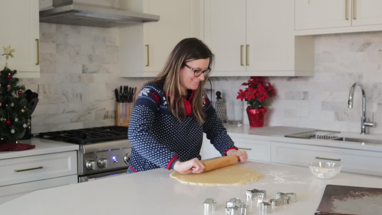 A happy woman rolls out cookie dough in a nice bright kitchen. Cookie cutters for Christmas cookies are ready to be used. Festive holiday decor in the background