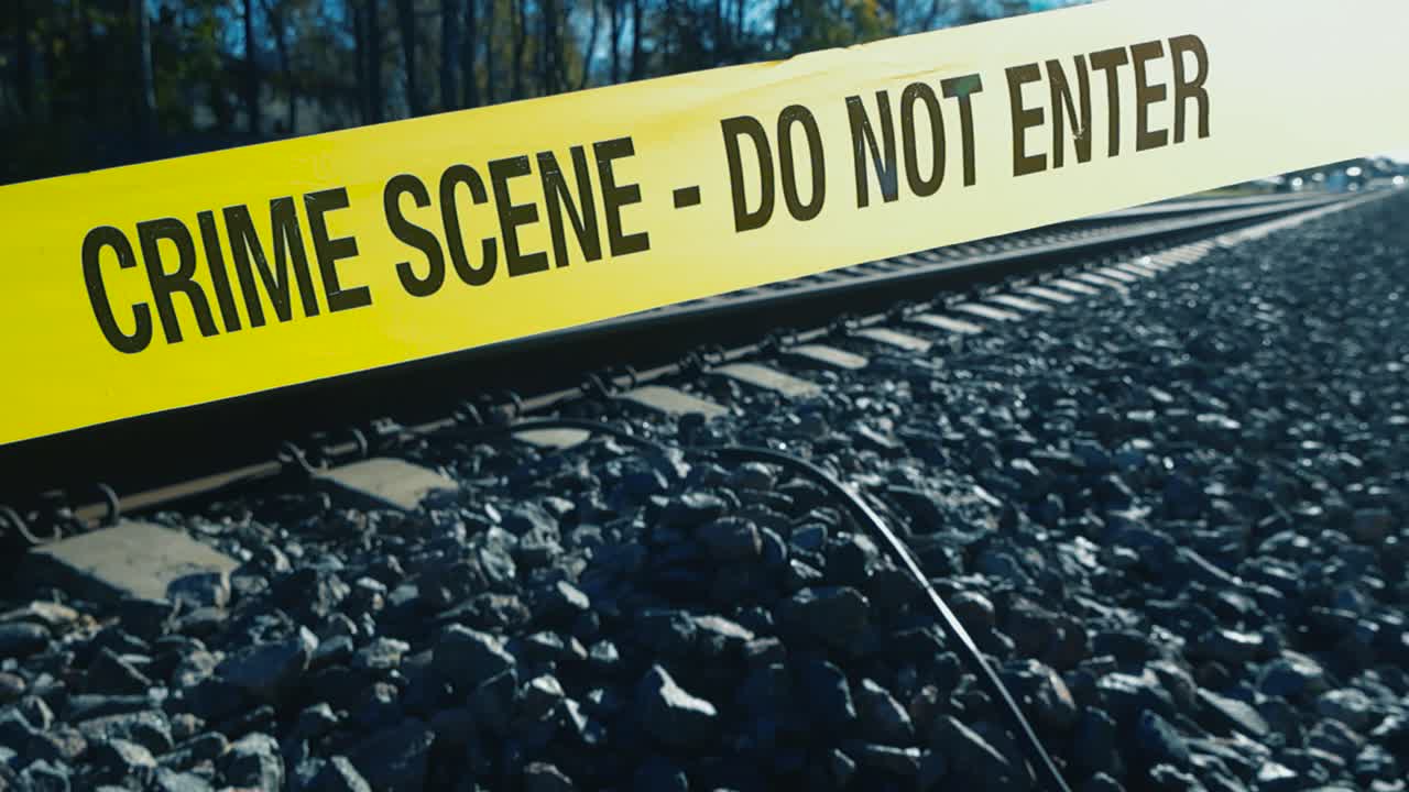 Yellow police line crime scene forensic investigation tape ribbon in front of railroad or train tracks during a sunny day with sunlight backlighting the tape. Gravel and grass around the train tracks