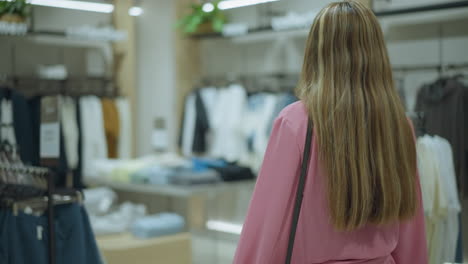 back view of a lady in pink cloth and black hand bag walking through a clothes rack in a well-lit shopping mall, clothes are hung around, with a blurred view of other items in the background