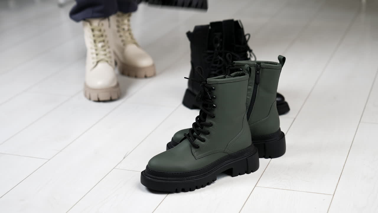 Two pairs of black and grey boots on tractor soles on the floor. Woman in white boots comes up bringing beige and black footwear.