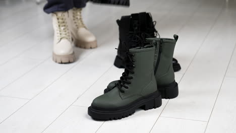 two pairs of black and grey boots on tractor soles on the floor. woman in white boots comes up bringing beige and black footwear.