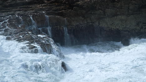 Wave-crash-into-rocks,-frothing-sea-water-streams-down-rock-face,-turbulent-ocean