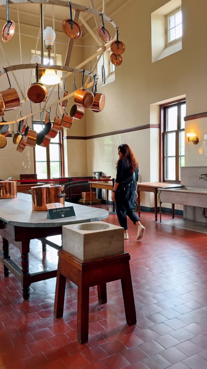 Cucina di una dimora storica con pentole di rame appese e un visitatore. Ampia cucina caratterizzata da pentole di rame appese, un tavolo da lavoro centrale e una persona che attraversa la stanza.