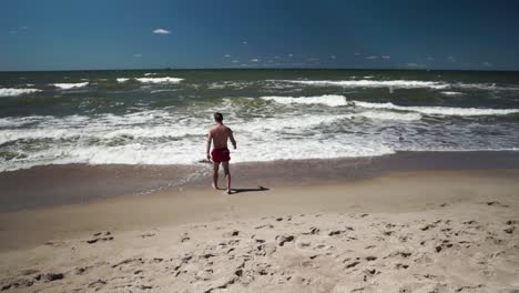 Junger-Mann-In-Roten-Shorts-Geht-An-Einem-Strand-Ins-Meer-1