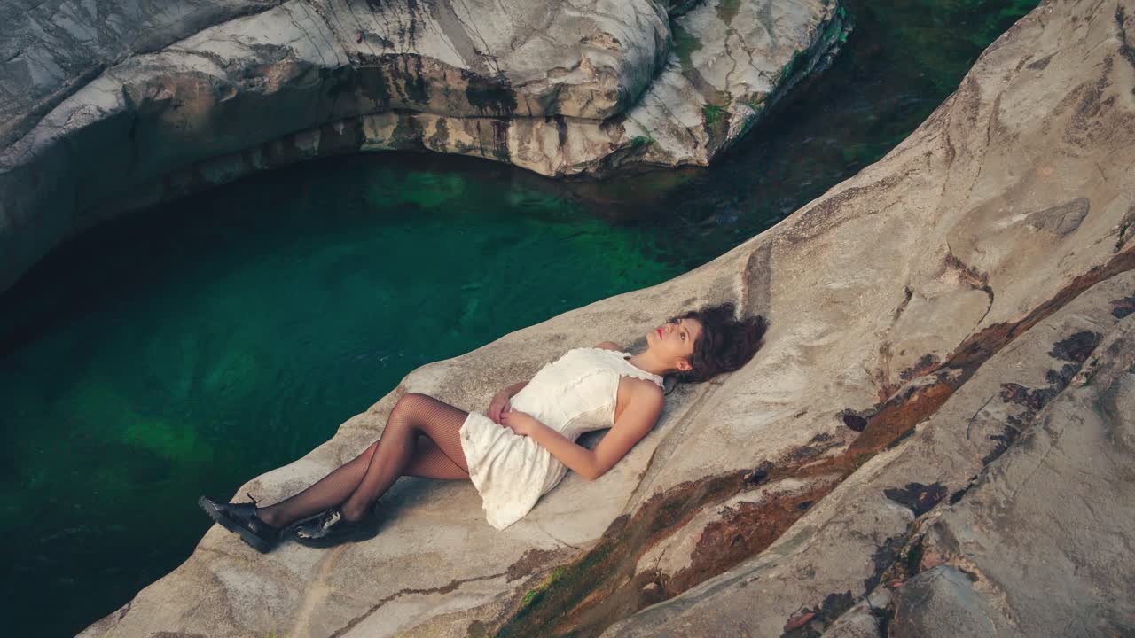 Cinemagraph - seamless video loop of a young brunette model girl in a white dress sitting on the edge of a gorge canyon by a blue mountain river. The water is rushing through naturally formed rocks.