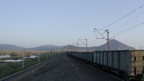 Toma-Aérea-De-Seguimiento-De-Un-Tren-Largo-De-Carruajes-De-Carbón-Completamente-Cargado,-Que-Pasa-Por-Campos-Verdes,-Con-La-Cresta-De-La-Montaña-A-Lo-Lejos,-Al-Atardecer