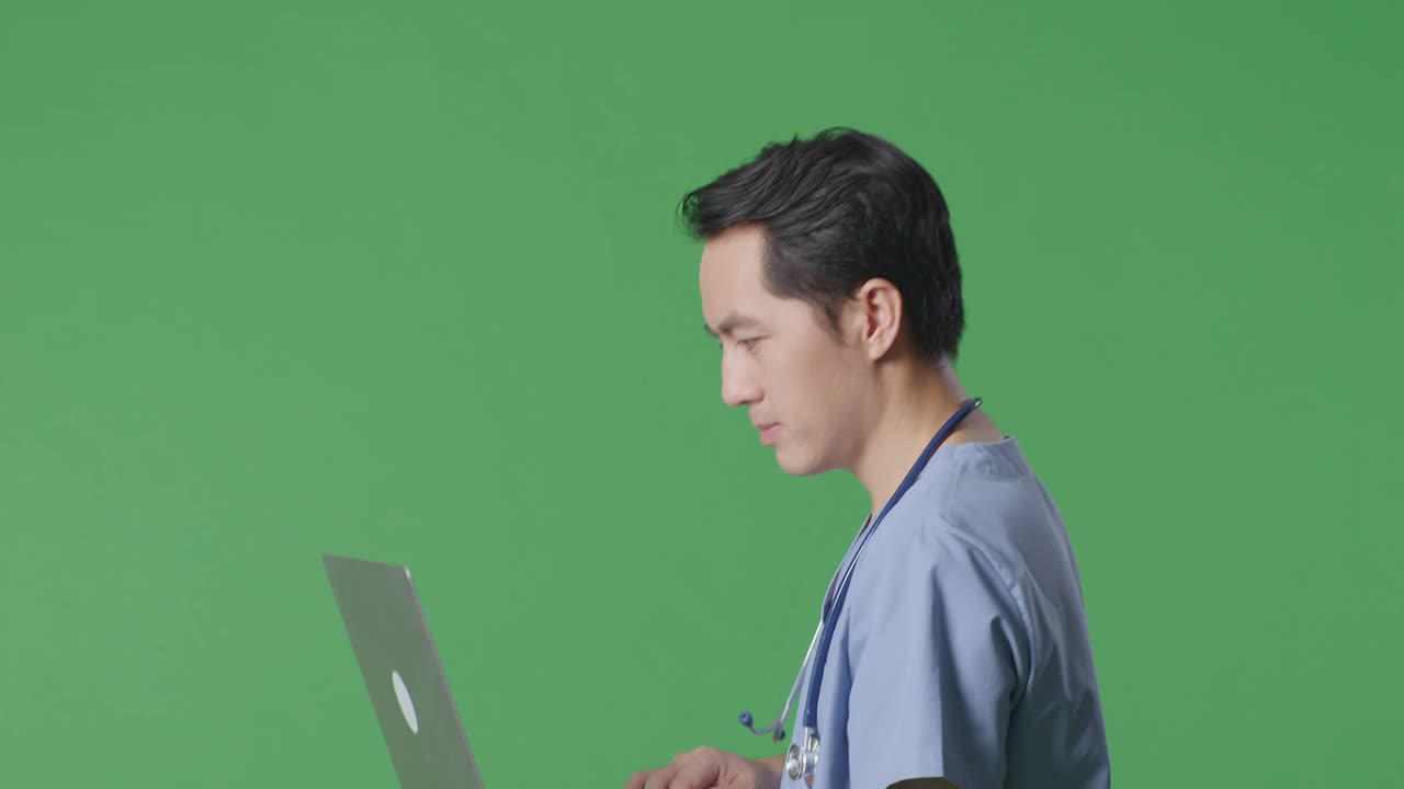 병원 에서 녹색 스크린 배경 에 걸어 다니는 동안 노트북 을 사용 하는 스테토스코프 를 들고 있는 아시아 남성 의사