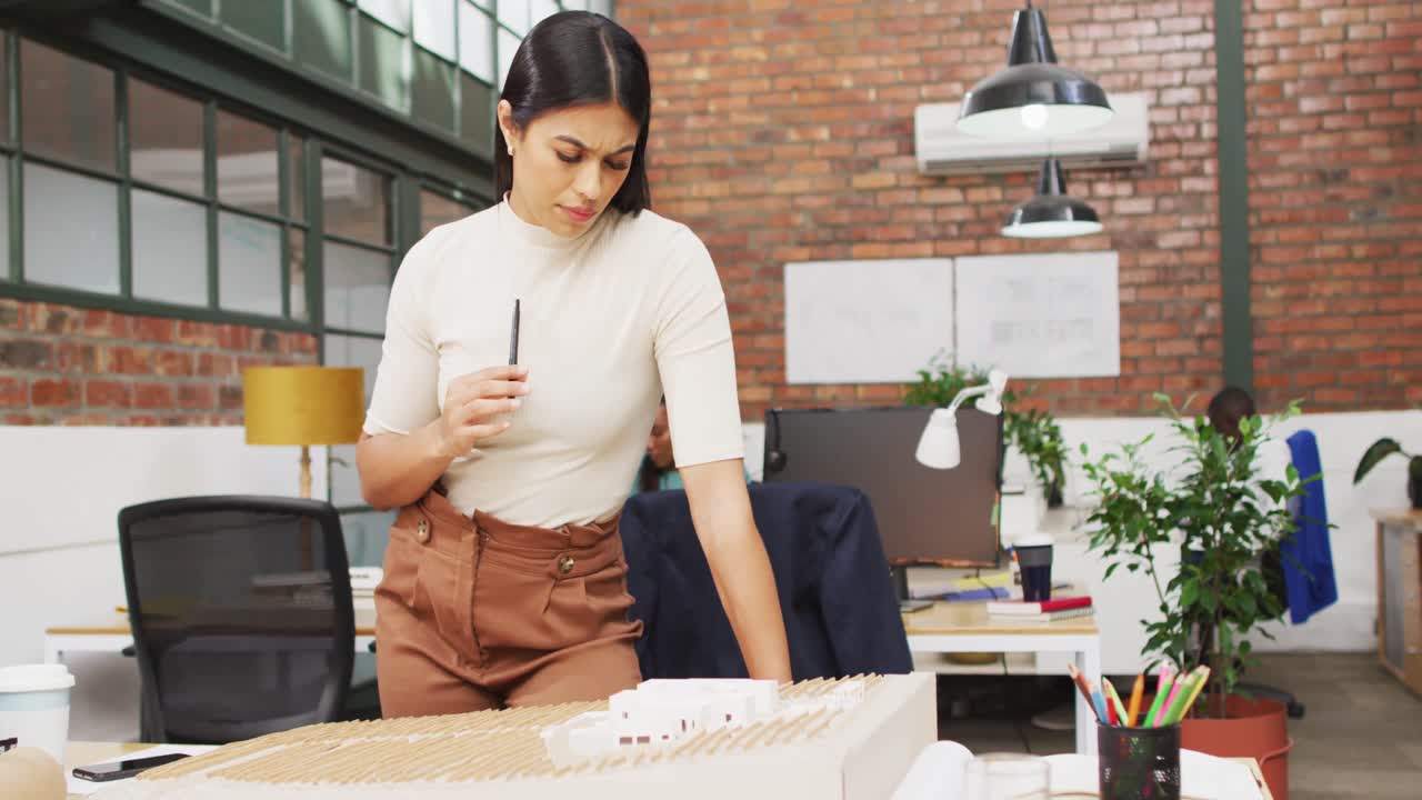 arquitecta biracial pensativa que mira modelos arquitectónicos en la oficina