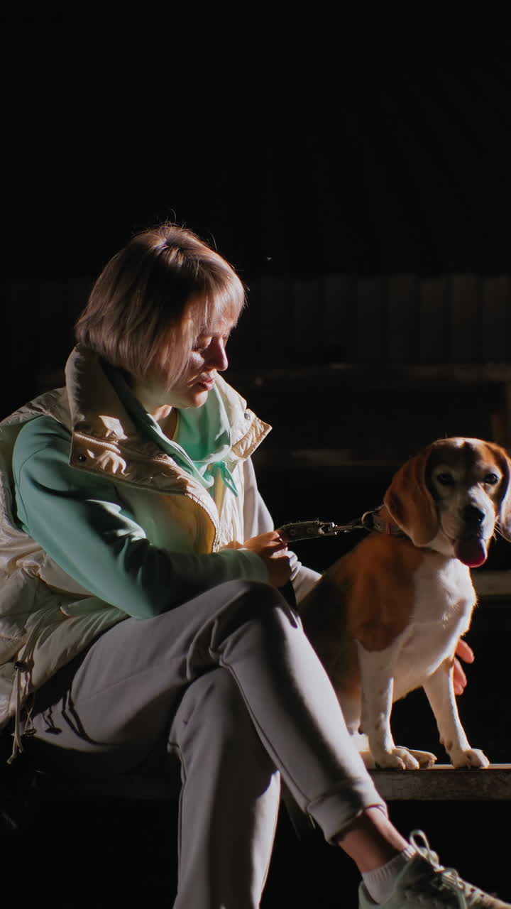 흰색 옷을 입은 여성과 비글이 따뜻한 측면광 아래 어두운 관람석에서 교감하는 모습, 민트색 조끼와 운동화, 명령과 간식으로 하는 즐거운 훈련 세션, 주의 깊고 기민한 강아지, 클로즈업 인물 사진