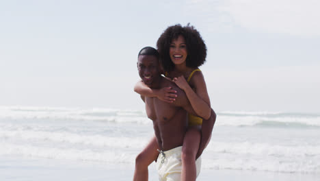 una pareja afroamericana sonriente jugando en la playa, un hombre llevando a una mujer a lomos